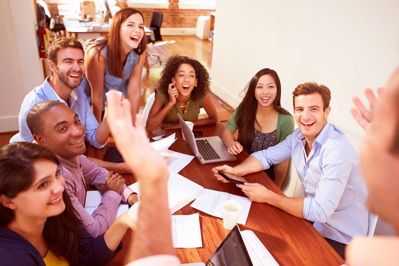 A group of young mutli-ethnic copywriters sitting around a table, with their laptops, and raising their hands at a Quantico Copywriting Course in Singapore